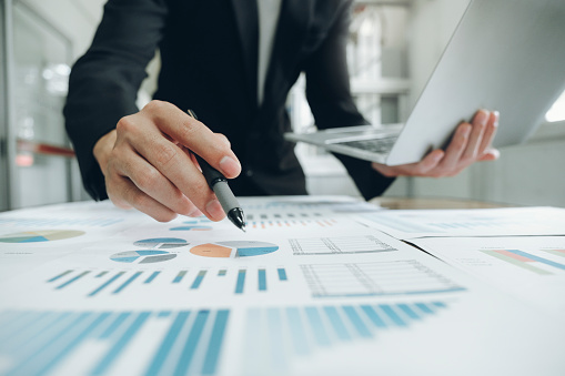 Close up of hand holding pen, hovering over a graph, while the other hand holds an open laptop.